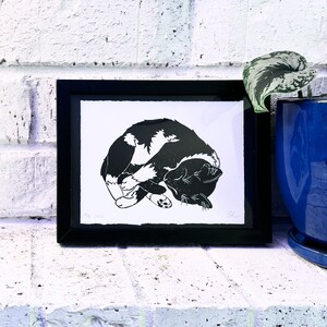 The print framed in a black frame and sitting on a white brick mantle. A green pothos plant in a dark blue pot sits next to the frame.
