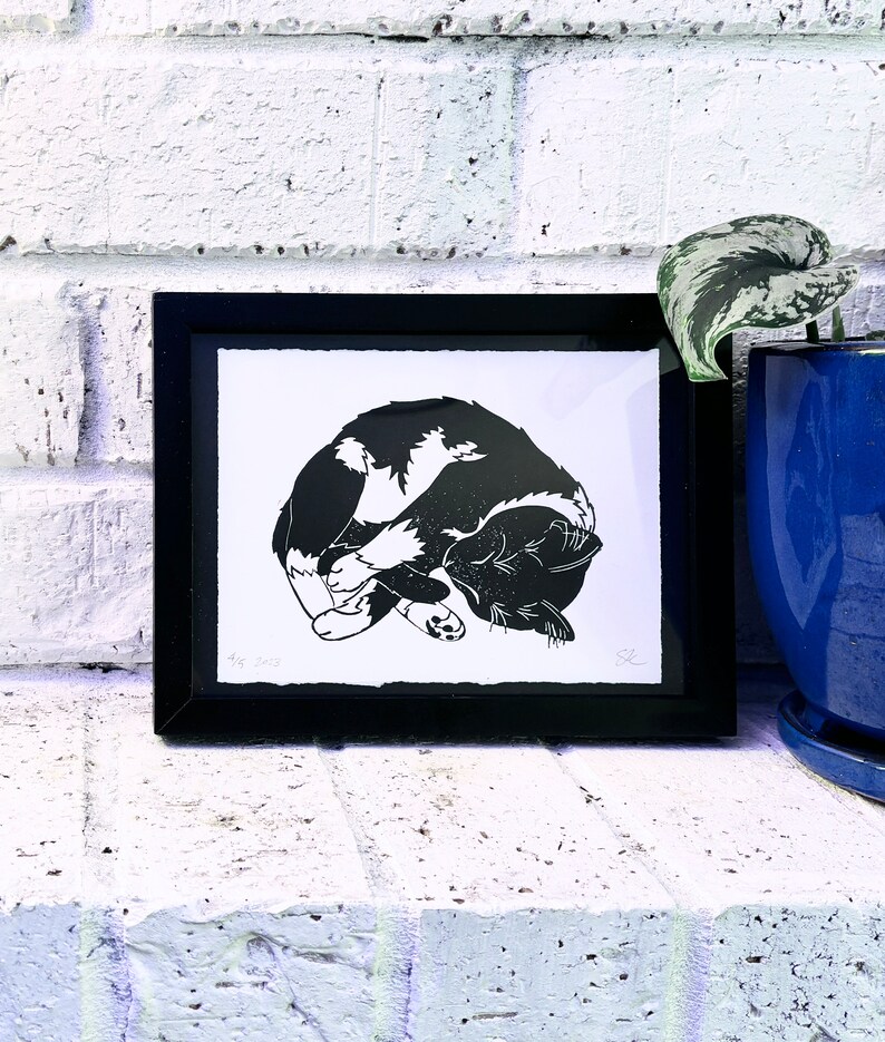The print framed in a black frame and sitting on a white brick mantle. A green pothos plant in a dark blue pot sits next to the frame.