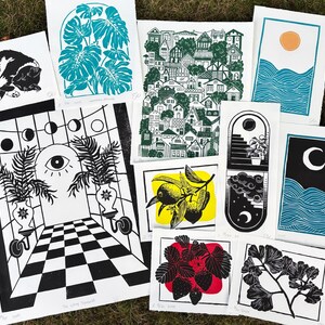 Some of the artists other prints. A pair of sun and moon over the blue ocean prints, a print with about fifty houses stacked as though on a hill, a leafy monstera, a sleeping tuxedo cat, a ginkgo branch, lemon branch, and strawberry plant. there is also a large print of a magical portal archway with an eye on it and checkerboard floor leading up to it, and two large leafy potted plants on either side.