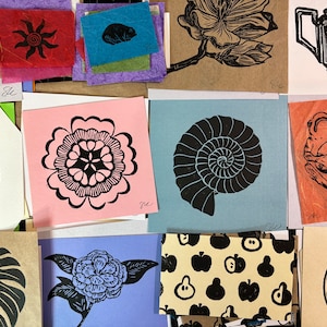May include: A collection of 10 small, colorful cards with black and white linocut prints of various designs, including a flower, a nautilus shell, a frog playing a guitar, a sun, a ginkgo leaf, a monstera leaf, a citrus fruit, and a flower with leaves.