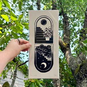The print packaged in a clear plastic sleeve with a sheet of cardboard behind it. It is being held in front of green tree branches.