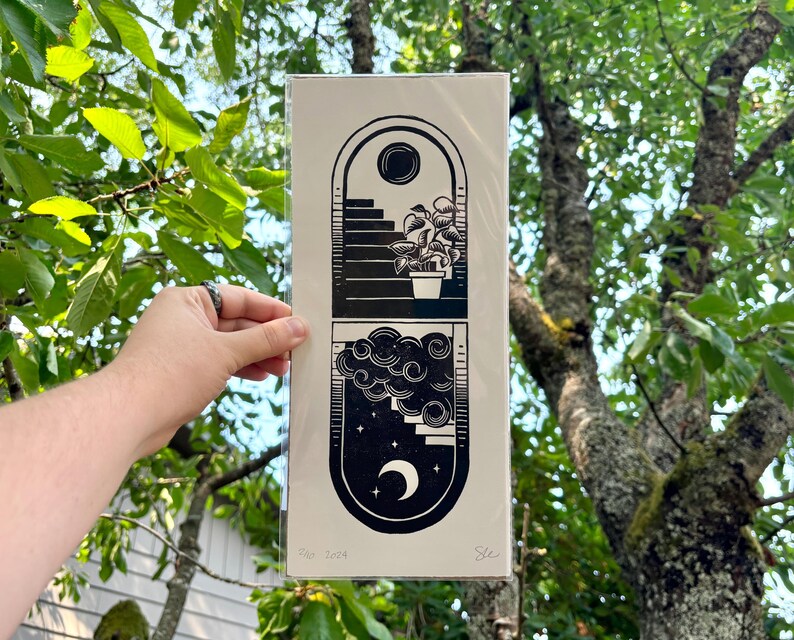 The print packaged in a clear plastic sleeve with a sheet of cardboard behind it. It is being held in front of green tree branches.