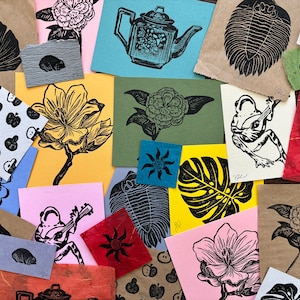 May include: A collection of handmade paper prints featuring various designs, including a ginkgo leaf, a flower, a trilobite, a frog, and a monstera leaf. The prints are on different colored papers and have a rustic, hand-crafted aesthetic.