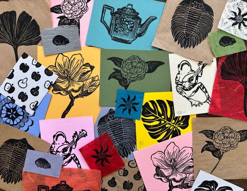 May include: A collection of handmade paper prints featuring various designs, including a ginkgo leaf, a flower, a trilobite, a frog, and a monstera leaf. The prints are on different colored papers and have a rustic, hand-crafted aesthetic.