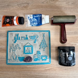 The linoleum block, ink, and rollers used to make this print. The block is a reverse image of the print. There is a pink rubber cat stamp sitting on the linoleum in the same place where the cat image is printed in the art piece.