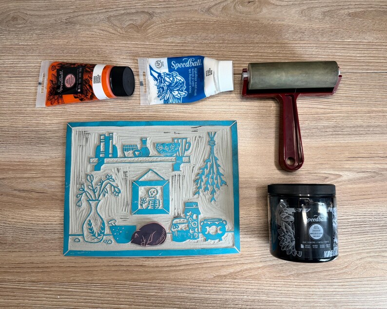 The linoleum block, ink, and rollers used to make this print. The block is a reverse image of the print. There is a pink rubber cat stamp sitting on the linoleum in the same place where the cat image is printed in the art piece.