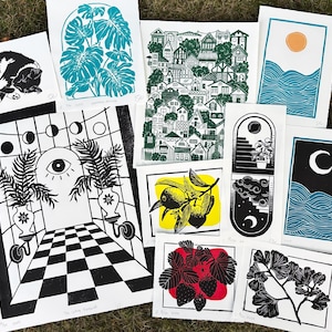 Some of the artists other prints. A pair of sun and moon over the blue ocean prints, a print with about fifty houses stacked as though on a hill, a leafy monstera, a sleeping tuxedo cat, a ginkgo branch, lemon branch, and strawberry plant. there is also a large print of a magical portal archway with an eye on it and checkerboard floor leading up to it, and two large leafy potted plants on either side.
