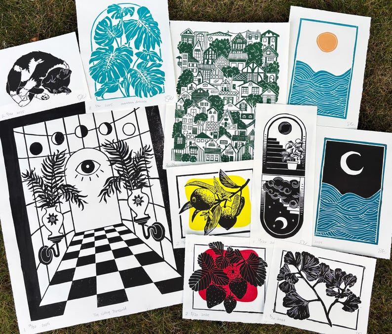 Some of the artists other prints. A pair of sun and moon over the blue ocean prints, a print with about fifty houses stacked as though on a hill, a leafy monstera, a sleeping tuxedo cat, a ginkgo branch, lemon branch, and strawberry plant. there is also a large print of a magical portal archway with an eye on it and checkerboard floor leading up to it, and two large leafy potted plants on either side.