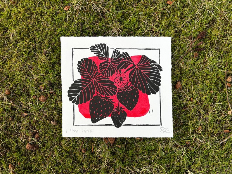 Horizontally oriented linocut print of a plant with three groups of black three leaves, three black strawberries hanging below, and one white flower in the center. A blobby red rectangle is behind the plant inside the border