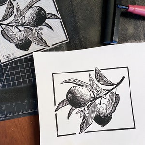 a process photo of the artist holding a lemon print with wider borders than the finished print has. there is the linoleum block used to print, which is an inverse image of the print. there is an ink roller on black ink, all on a craft mat.