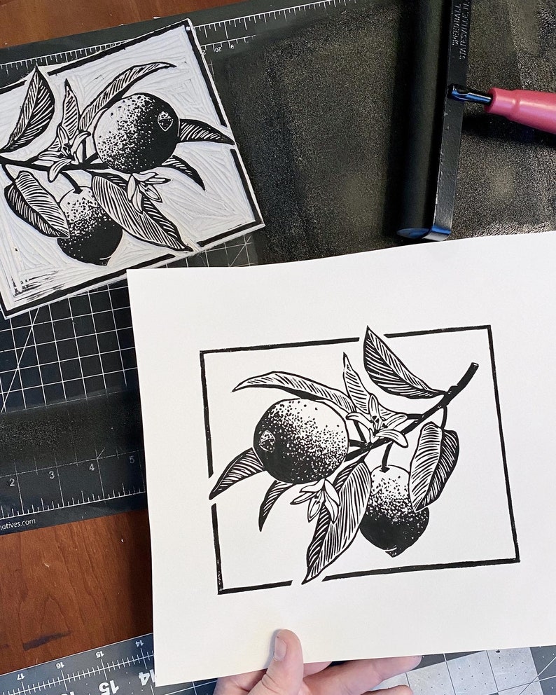 a process photo of the artist holding a lemon print with wider borders than the finished print has. there is the linoleum block used to print, which is an inverse image of the print. there is an ink roller on black ink, all on a craft mat.