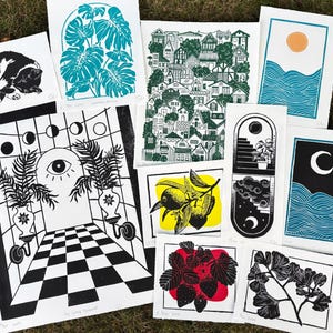Some of the artists other prints. A pair of sun and moon over the blue ocean prints, a print with about fifty houses stacked as though on a hill, a leafy monstera, a sleeping tuxedo cat, a ginkgo branch, lemon branch, and strawberry plant. there is also a large print of a magical portal archway with an eye on it and checkerboard floor leading up to it, and two large leafy potted plants on either side.