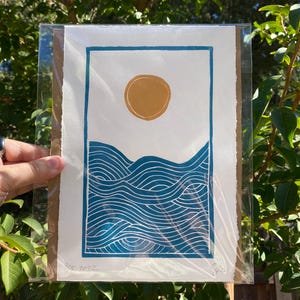 The print packaged in a clear plastic sleeve with a sheet of cardboard behind it. It is being held in front of green tree branches.