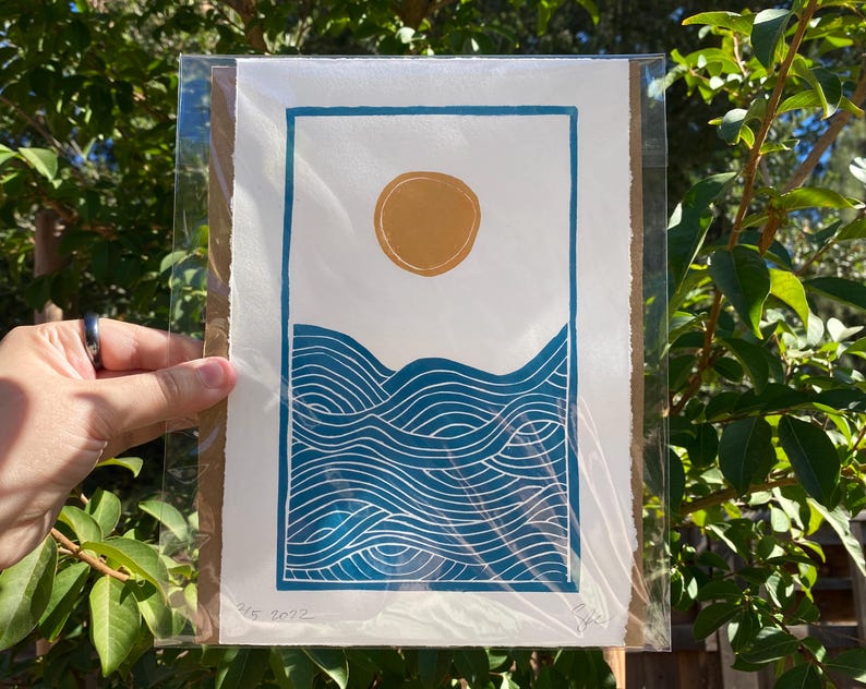 The print packaged in a clear plastic sleeve with a sheet of cardboard behind it. It is being held in front of green tree branches.