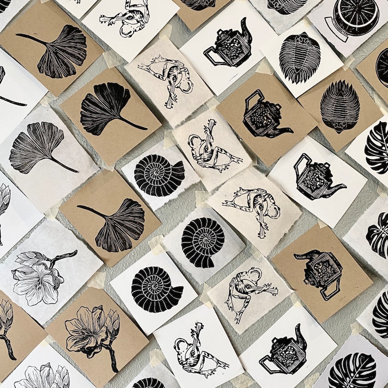 May include: A collection of black and white linocut prints featuring various designs, including leaves, flowers, teapots, and abstract shapes. The prints are arranged on a white surface.