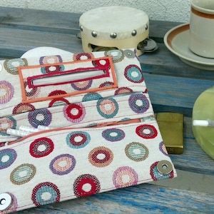 May include: A multi-colored fabric pouch with a zippered pocket and a snap closure. The pouch is decorated with a pattern of colorful circles. The pouch is open and there is a white cigarette inside.