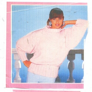 May include: A light pink, long-sleeved pullover sweater with a high neckline. The sweater has a relaxed fit and is paired with light-colored pants. The image is framed with a pink border and features the text "951/54 Fancy Pullover" and "Size Petite-Large".