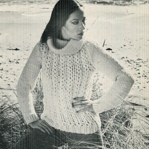May include: Cream-colored, chunky knit sweater with a high, ribbed turtleneck. The sweater has long sleeves and a textured, open knit pattern. The model is wearing the sweater on a beach with tall grass in the foreground.
