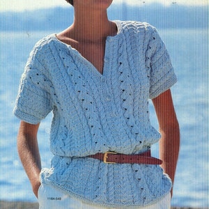 May include: A light blue knitted top with short sleeves, a V-neck, and a brown belt with a gold buckle. The top features a textured cable knit design and is labeled "Carmen". The model is wearing white pants.
