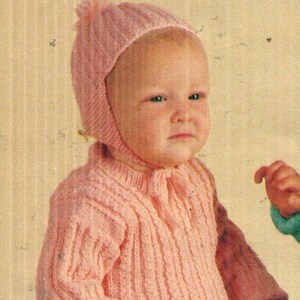 Könnte beinhalten: Ein hellrosa gestrickter Langarmpullover mit vertikaler Rippenstruktur und eine passende Strickmütze mit einem kleinen Bommel oben und einem Kinnband. Der Pullover hat einen Rundhalsausschnitt sowie gerippte Bündchen an Ärmeln und Taille.