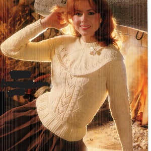 May include: Cream-colored knitted sweater with a decorative collar and intricate cable knit detailing. The sweater features a round neckline and long sleeves. A small, decorative pin is attached to the collar. The model is wearing a brown skirt.