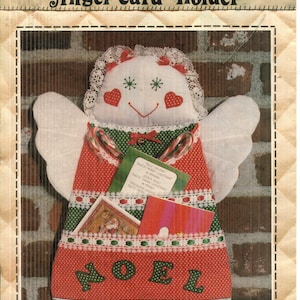 Może przedstawiać: Wzór rzemieślniczy zatytułowany Patch Press Angel Card Holder No. 322B. Obraz przedstawia materiałowego anioła z białą twarzą, koronkowym wykończeniem oraz czerwono-zieloną sukienkę w kropki z napisem NOEL i kieszeniami mieszczącymi kartki.
