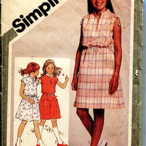 Puede incluir: Sobre de patrón de costura Simplicity vintage con un diseño de vestido a cuadros para niñas. El sobre incluye ilustraciones del vestido y el texto "Simplicity" en letras grandes y en negrita. El sobre está envejecido y desgastado.
