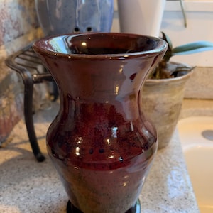 May include: A dark red ceramic vase with a glossy finish. The vase has a wide mouth and a narrow base.