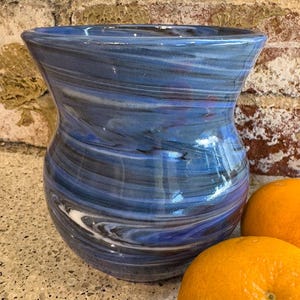 May include: A blue and white swirled glass vase with a wide opening and a rounded body. The vase has a glossy finish and is sitting next to two oranges. The vase is a decorative item.