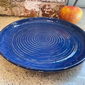 May include: A round, deep blue ceramic plate with concentric white grooves. The plate is on a speckled surface, with a red and yellow apple in the background. The plate is approximately 10 inches in diameter.