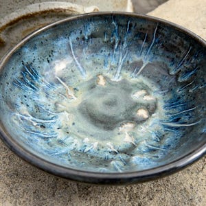 May include: A round ceramic bowl featuring a reactive glaze with deep navy blue, light blue, and grey tones. The interior surface displays a mottled, streaked pattern radiating from the center toward the dark, smooth, and rounded rim.