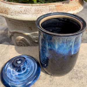 May include: A cylindrical ceramic canister with a deep blue and black glaze featuring a glossy finish and a matching removable lid with a central knob. The exterior surface displays a smooth texture with subtle color variations and drips.