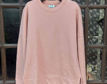 May include: A dusty pink long-sleeve sweatshirt with a crew neckline and dropped shoulders. The garment features a ribbed collar, cuffs, and hem, with small side slits at the bottom. A white label with the text Acne Studios is at the neck.