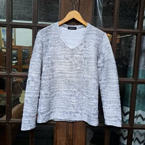 May include: A long-sleeved, gray V-neck shirt with a textured, horizontal striped pattern. The shirt is hanging on a wooden hanger. The fabric appears to be lightweight.