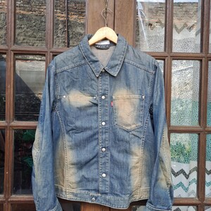 Puede incluir: Chaqueta vaquera azul con efecto lavado. La chaqueta tiene cierre de botones, cuello y bolsillo en el pecho con etiqueta roja. La chaqueta está colgada de una percha de madera.