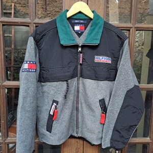 May include: A gray, black, and green Tommy Hilfiger zip-up jacket. The jacket has a green collar, black chest panels, and gray sleeves. The Tommy Hilfiger logo is visible on the left sleeve and chest. The jacket is hanging on a wooden hanger.