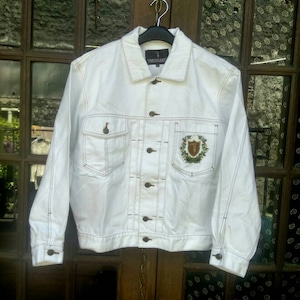 May include: White denim jacket with brown stitching and a small embroidered logo on the left chest that says "THUSSAUD".