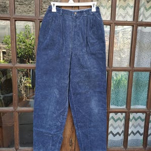 Könnte beinhalten: Hoch taillierte Cordhose in tiefem Marineblau. Diese Hose verfügt über ein plissiertes Vorderteil, einen Verschluss mit Knopf und Reißverschluss sowie Gürtelschlaufen am Bund. Der Stoff weist eine ausgeprägte vertikale Rippenstruktur auf.