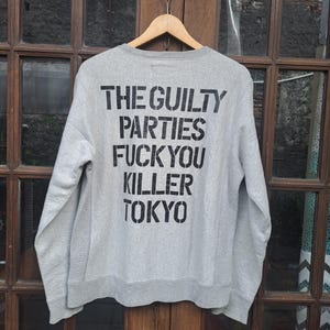 May include: A light gray sweatshirt with the text "THE GUILTY PARTIES FUCK YOU KILLER TOKYO" printed in black. The sweatshirt is hanging on a wooden hanger against a window.