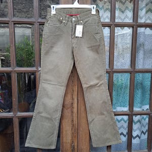May include: Olive green denim pants hanging on a white hanger. The pants have a classic five-pocket design, a button and zipper closure, and a red interior waistband. A price tag hangs from the belt loop.
