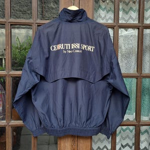 Könnte beinhalten: Marineblaue Windjacke mit hohem Kragen und elastischem Saum. Auf der Rückseite der Jacke steht in weißen Buchstaben "CERUTTI 1881 SPORT by Nino Cerruti". Die Jacke hängt an einem Holzkleiderbügel.