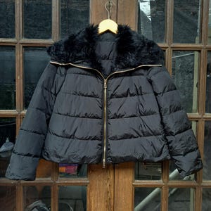 Puede incluir: Una chaqueta acolchada negra con cuello de piel sintética. La chaqueta tiene cierre de cremallera y un diseño acolchado.