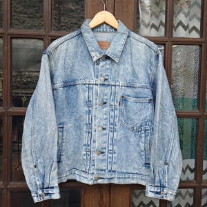 Op de afbeelding: Een lichtblauw denim jack met een verbleekte look. Het jack heeft een knoopsluiting, twee borstzakken en twee voorzakken. Het jack hangt aan een houten hanger.