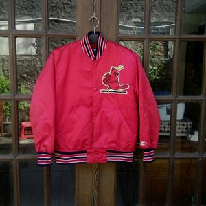 May include: A red satin baseball jacket with a St. Louis Cardinals logo on the front. The jacket has a ribbed collar, cuffs, and waistband. The logo is a red cardinal bird with a baseball bat.