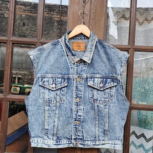 Könnte beinhalten: Eine ärmellose Jeansjacke in hellblauer Waschung, die an einem Holzbügel hängt. Die Jacke hat einen Kragen, eine Knopfleiste vorne und zwei Brusttaschen. Ein brauner Lederaufnäher ist auf dem Kragenrücken sichtbar.