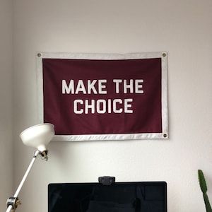 May include: A rectangular maroon fabric wall banner with a white border and metal grommets in the corners. The text "MAKE THE CHOICE" is printed in large, bold, white capital letters across the center of the maroon field of the banner.