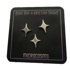 Może przedstawiać: Trzy srebrne szpilki w kształcie gwiazdy na czarnym kartoniku z tekstem "Born Rad x Kristina Drake" i "Microcosms".