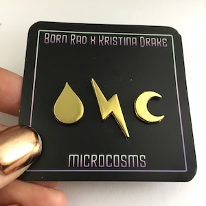 May include: Gold-colored pins with a water drop, lightning bolt, and crescent moon design. The pins are on a black card with the text "Born Rad x Kristina Drake" and "Microcosms".