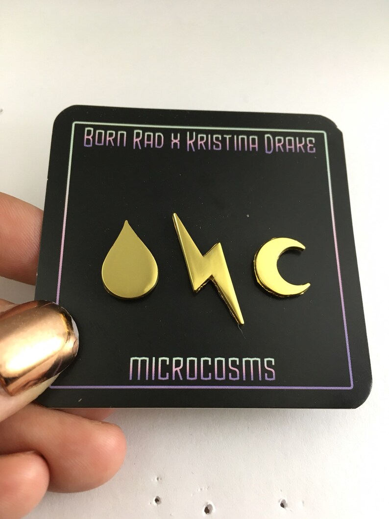 May include: Gold-colored pins with a water drop, lightning bolt, and crescent moon design. The pins are on a black card with the text "Born Rad x Kristina Drake" and "Microcosms".
