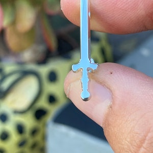 May include: A silver sword pin with a detailed design. The pin is held in a hand.